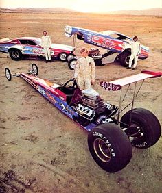 two men standing next to a race car in the desert with other cars behind them