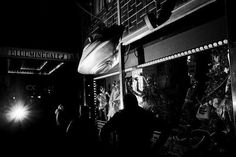 black and white photograph of people walking in front of a store at night with lights on