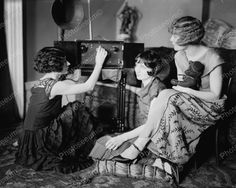 three women sitting on the floor in front of an old fashioned radio set, with one woman reaching up for something