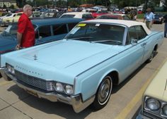 an old blue car parked in a parking lot next to other older cars and people