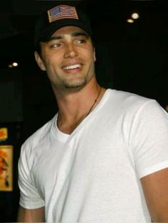 a man in a white t - shirt and black hat smiles at the camera while holding a cell phone