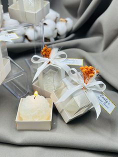 several small white boxes with candles inside on a gray cloth covered tablecloth, next to cotton balls and an orange flower