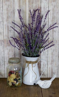 a white vase filled with purple flowers next to a jar full of coins and a bird figurine