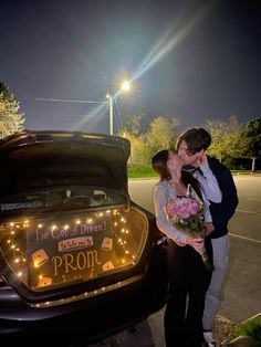 a man and woman kissing in front of a car with its trunk open at night