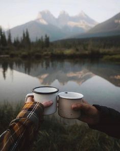 two people holding coffee cups in their hands near a lake with mountains in the background