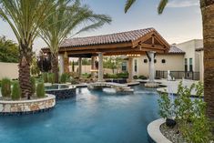 an outdoor swimming pool surrounded by palm trees and other landscaping items in front of a house