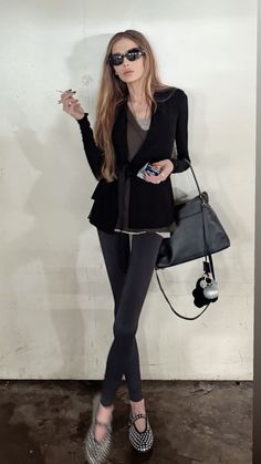 a woman in black jacket and leggings holding a handbag while standing next to a wall