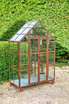 a small wooden greenhouse sitting on top of a gravel ground next to a green hedge