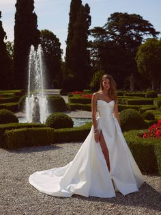 a woman standing in front of a fountain wearing a white wedding dress with high slit