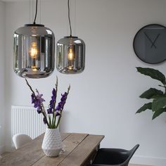 a wooden table topped with two vases filled with purple flowers next to a wall mounted clock