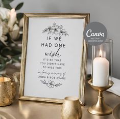 a white and gold framed sign with a candle next to it on a table in front of flowers