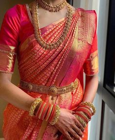 a woman in an orange and pink saree with gold jewelry on her neck, standing next to a window