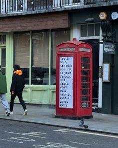 two people walking past a red phone booth on the side of the road in front of a building