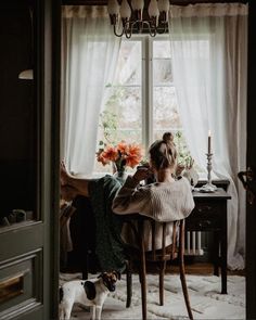 a woman sitting at a table in front of a window next to a small dog