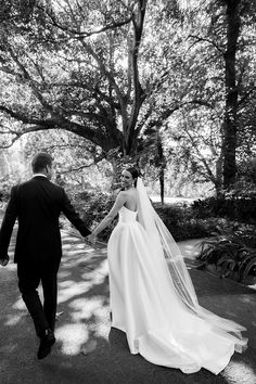 a bride and groom holding hands walking through the park in black and white, with trees behind them