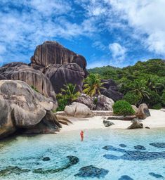 a person standing in the water near large rocks and boulders on an island with palm trees