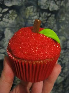 a hand holding a red cupcake with a green leaf on top