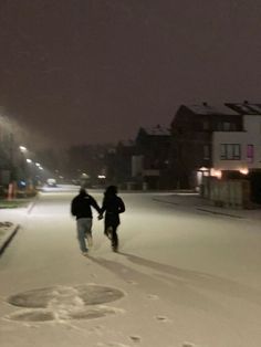 two people are running in the snow on a street at night with buildings behind them