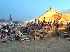 many people are sitting on wooden pallets in front of a large tent at night