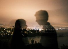 two people standing next to each other in front of a city at night with the lights on