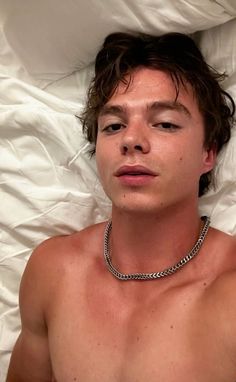 a shirtless young man laying in bed wearing a chain around his neck and chest