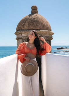 a woman in an orange top and white pants standing on a balcony next to the ocean