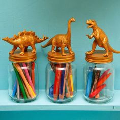 three glass jars filled with different colored pencils and plastic dinosaur figurines on top of a shelf