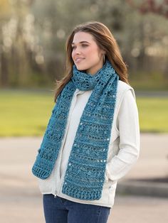 a woman wearing a blue crochet scarf standing in front of a park with trees