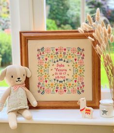 a teddy bear sitting next to a framed cross - stitch pattern on a window sill