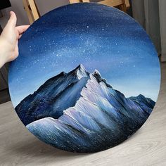 a hand holding up a painting of a mountain range under the stars in the night sky