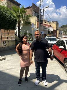 a man and woman standing next to each other in front of parked cars on a city street