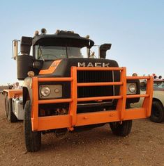 an orange mack truck parked in the middle of a dirt field with other trucks behind it