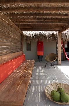 a wooden bench sitting under a thatched roof next to two green apples and a red surfboard