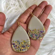 a pair of white earrings with gold and multicolored crystals on them, in the palm of someone's hand