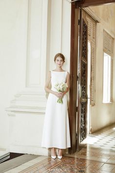 a woman in a white dress holding a bouquet standing next to an open door and looking at the camera