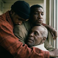 two men are hugging each other in front of a window and one is wearing a red sweater