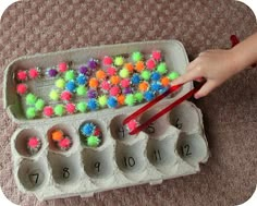 an egg carton filled with lots of colorful pom - poms next to a child's hand holding a pair of red chopsticks