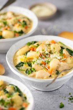 three white bowls filled with soup on top of a gray table next to bread and spoons