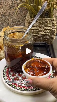 a person is holding a spoon over a jar of jam on a plate next to a potted plant