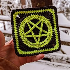 a hand holding up a crocheted square with a pentagram on it in front of snow covered trees