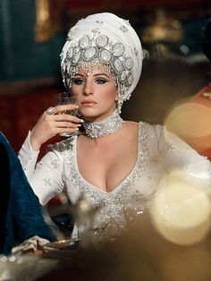 a woman in a white dress and headdress sitting at a table with food