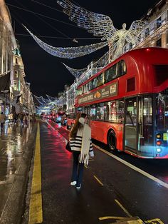 a red double decker bus driving down a street next to tall buildings covered in christmas lights