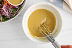 a hand holding a whisk over a bowl filled with dressing next to a salad