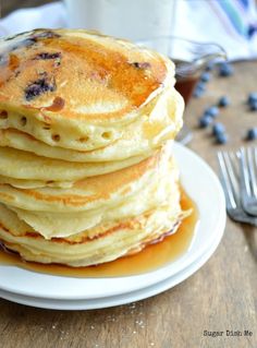 a stack of pancakes on a white plate with syrup and blueberries next to it