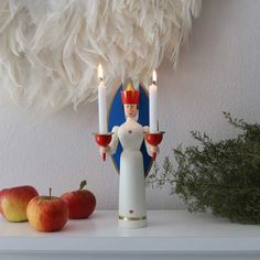 a white figurine holding two candles on top of a mantle next to apples