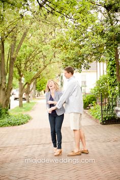 a man and woman holding hands while walking down a brick path in front of trees