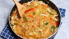 a wok filled with noodles and vegetables on top of a blue towel next to a wooden spoon