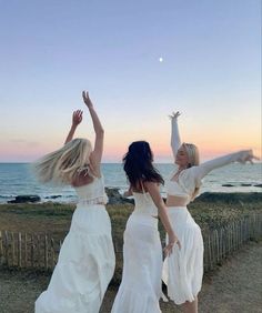 two women in white dresses are dancing near the ocean at sunset with their arms up