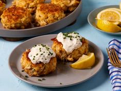 two plates with crab cakes and lemon wedges on blue table cloth next to silverware