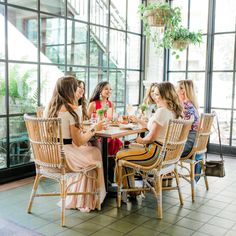 a group of women sitting around a wooden table eating food and drinking wine in front of large windows